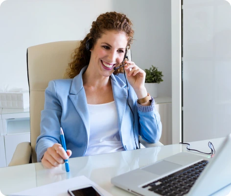 A woman wearing a headset and a light blue blazer smiles while sitting at a desk and looking at her laptop. She holds a pen and appears to be engaged in a video call or virtual meeting with Letting Agents.